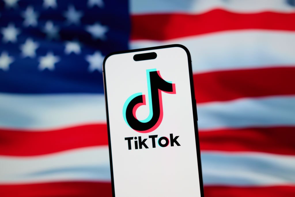 The TikTok app and logo are seen on a mobile device in front of a US flag